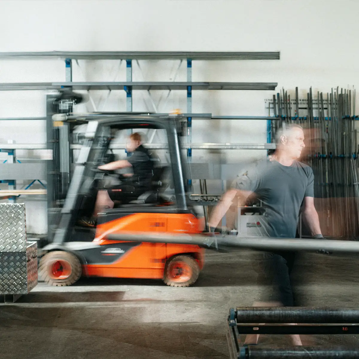 Arbeiter trägt Metallröhre in einer belebten Lagerhalle.